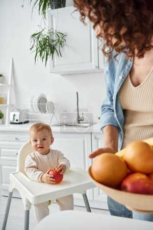 joyful child sitting in baby chair with ripe apple near mom with fresh fruits on blurred foreground