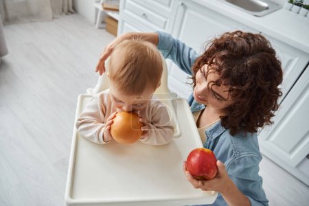 top view of little child in baby chair biting whole orange near smiling mother with ripe apple