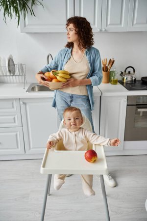 mujer pensativa con frutas frescas mirando hacia otro lado cerca de niño juguetón sentado en silla de bebé cerca de manzana