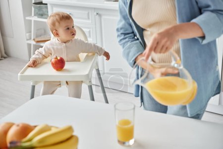 woman pouring fresh orange juice while toddler daughter sitting in baby chair near ripe apple