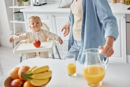 cute kid in baby char looking at ripe apple near mother with jug of orange juice, morning in kitchen