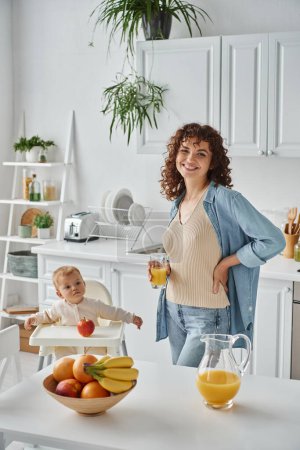 cheerful woman with orange juice standing with hand on hip near kid in baby chair and ripe fruits