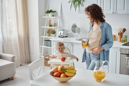 curly woman with glass of orange juice looking at child in baby chair in kitchen with ripe fruits