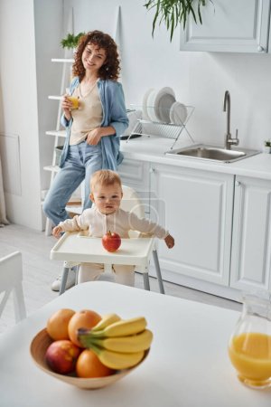 fröhliche Frau mit einem Glas Orangensaft neben dem Kleinkind im Babystuhl und reifen Früchten in der Küche