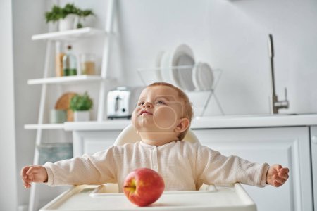 toddler girl sitting in baby chair near ripe apple and looking away in kitchen, happy childhood