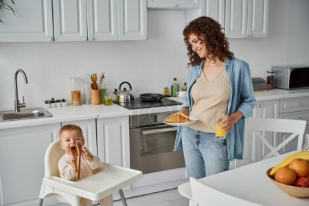 smiling woman with croissant and orange juice looking at funny child chewing wooden tongs in kitchen