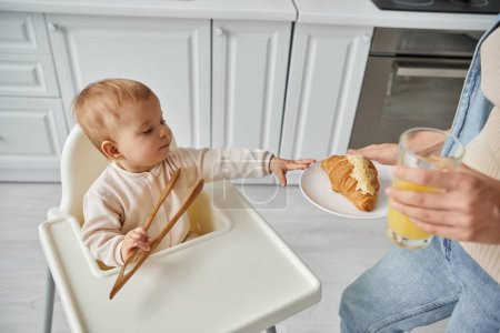 woman with croissant and fresh orange juice near toddler kid with wooden tongs in kitchen, breakfast