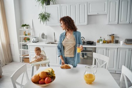 Frau mit Orangensaft und Croissant in Obstnähe und Kind spielt mit Holzzange in Küche