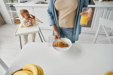 woman holding fresh orange juice and croissant near toddler daughter playing with wooden tongs