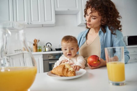 madre y bebé cerca de croissant, manzana madura y jugo de naranja fresco en la cocina, delicioso desayuno