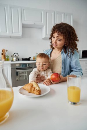 cute child reaching tasty croissant near fresh orange juice and mom with apple in kitchen, mealtime