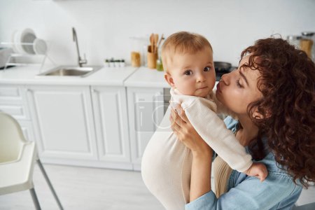 curly woman embracing and kissing toddler daughter in romper in cozy kitchen at home, family bliss