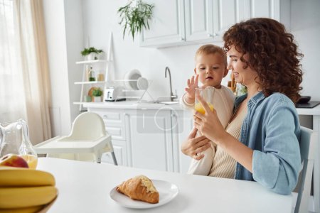 happy mother with baby and glass of orange juice near croissant and fruits in kitchen, breakfast