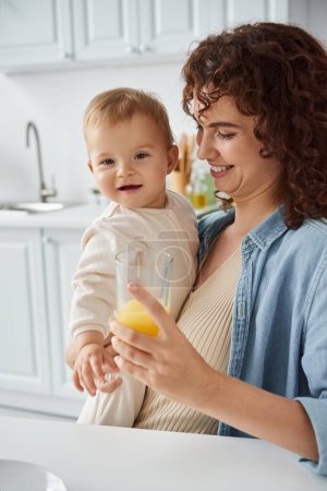 smiling woman holding glass of natural orange juice near carefree baby girl, happy family morning