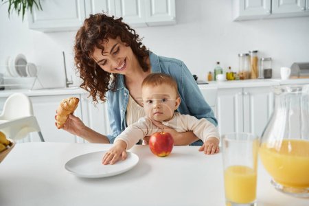 pleased woman holding tasty croissant near baby and apple with orange juice, morning mealtime