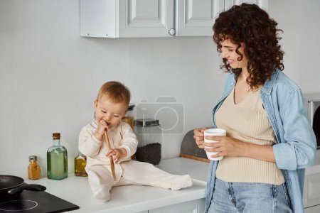 smiling woman with cup of coffee near cute child sitting on kitchen counter and chewing wooden fork