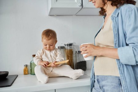 toddler girl in romper playing with wooden fork on kitchen worktop near mom with coffee cup