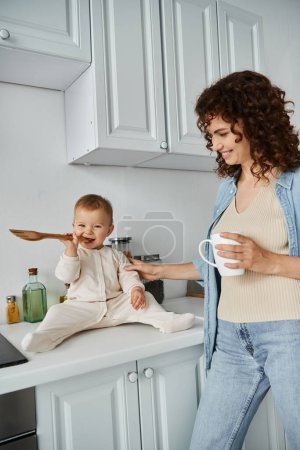 carefree baby sitting on kitchen counter and chewing wooden fork near smiling mother with coffee cup