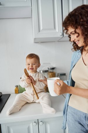 niño alegre sentado en la encimera de la cocina y jugando con tenedor de madera cerca de mamá feliz con taza de café