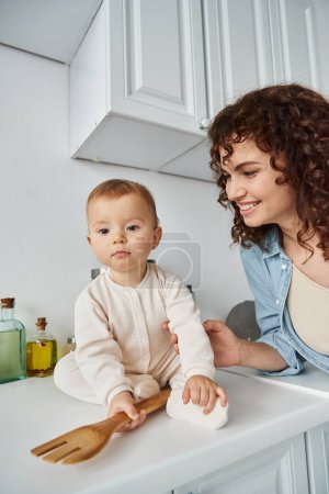 curly woman smiling near toddler daughter with wooden fork sitting on kitchen counter, morning fun