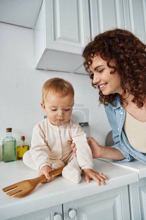 cheerful woman near little daughter with wooden fork sitting on kitchen worktop, happy morning