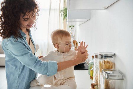 happy woman holding tiny hand of toddler daughter sitting on kitchen counter, blissful parenthood