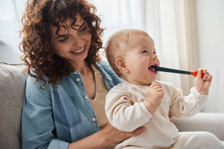 smiling woman sitting on couch with toddler daughter chewing teething toy in living room, playtime