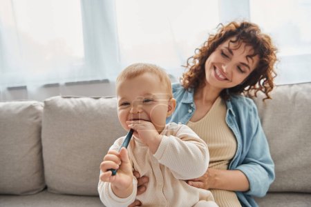 verspieltes Baby kaut Kinderspielzeug neben lächelnder Mutter auf Couch im Wohnzimmer, Qualitätszeit zu Hause