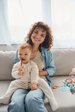 lockige Frau lächelt in die Kamera in der Nähe von Kleinkind Tochter kaut Kinderspielzeug auf Couch im Wohnzimmer