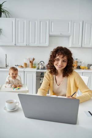 smiling woman working on laptop near coffee cup and toddler daughter on background in kitchen