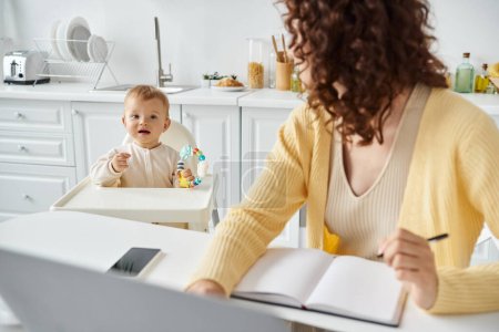girl with rattle toy looking at mom working near laptop and smartphone in kitchen, work life balance