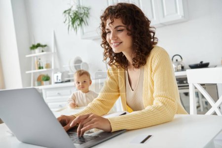 lächelnde lockige Frau tippt auf Laptop, während sie aus der Ferne in der Küche in der Nähe von Kleinkind-Tochter arbeitet