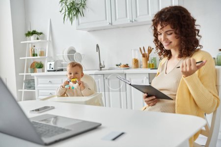 glückliche Frau sitzt mit Notizbuch neben Laptop und Kleinkind mit Rasselspielzeug in gemütlicher Küche