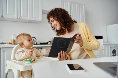 Foto de Mujer alegre mostrando cuaderno a la pequeña hija sentada en silla de bebé en la cocina, padre que trabaja - Imagen libre de derechos