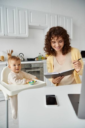 smiling woman looking in notebook near little child in baby chair in kitchen, career and parenthood