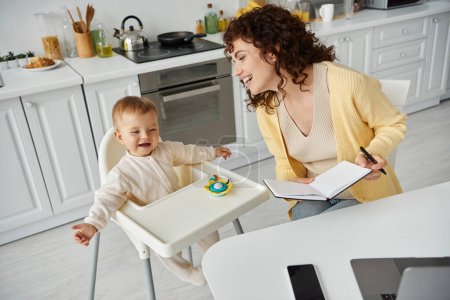 joyful mother with notebook near excited child sitting in baby chair with rattle toy, working parent