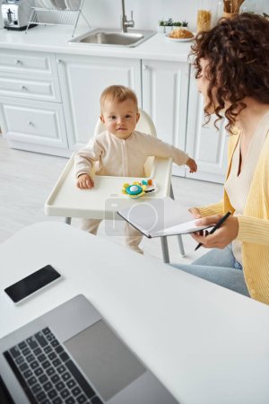niedliches Kind im Babystuhl neben der Mutter, die mit Notebook und Laptop in der Küche arbeitet, Work Life Balance
