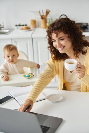 Photo for Female freelancer with coffee cup working on laptop near toddler child in kitchen, multitasking - Royalty Free Image