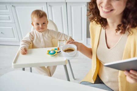 mujer con café de la mañana y portátil cerca de niño sonriente en silla de bebé con juguete sonajero en la cocina