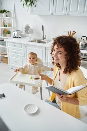 Lächelnde Frau mit Morgenkaffee und Notizbuch neben fröhlichem Kind, das im Kinderstuhl in der Küche sitzt
