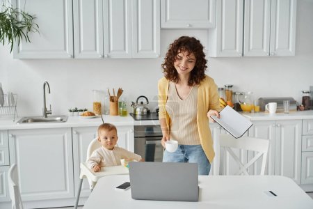 happy woman with notebook and coffee cup near laptop and toddler daughter in baby chair in kitchen