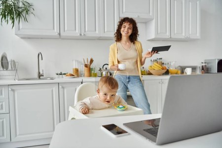 happy female freelancer with coffee cup and notebook near child in baby chair and devices in kitchen