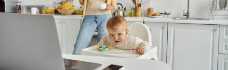 Foto de Niña sentada en silla de bebé en la cocina cerca de mamá con taza de café y computadora portátil en la mesa, pancarta - Imagen libre de derechos