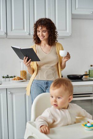 Photo for Smiling woman with coffee cup looking in notebook near toddler daughter in baby chair, multitasking - Royalty Free Image