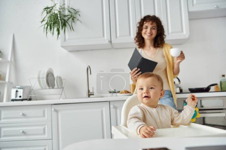 toddler child playing with rattle toy near mother with notebook and coffee cup in modern kitchen