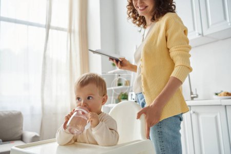 Foto de Niña sentada en silla de bebé y bebiendo agua cerca de la madre sonriente con cuaderno en la cocina - Imagen libre de derechos