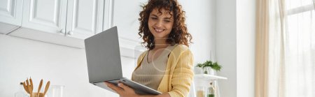 femme heureuse avec les cheveux ondulés debout avec ordinateur portable dans la cuisine moderne, pigiste et femme au foyer, bannière
