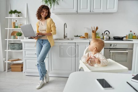 Photo for Mother with laptop looking at toddler daughter sitting with baby bottle in kitchen, work at home - Royalty Free Image