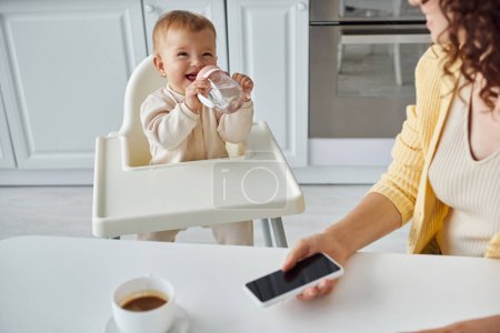 fröhliches Kind trinkt aus Babyflasche in der Nähe der Mutter mit Smartphone mit leerem Bildschirm in der Nähe der Kaffeetasse