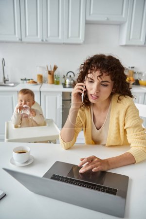 Photo for Busy woman talking on smartphone and working on laptop near little child in kitchen, freelance work - Royalty Free Image